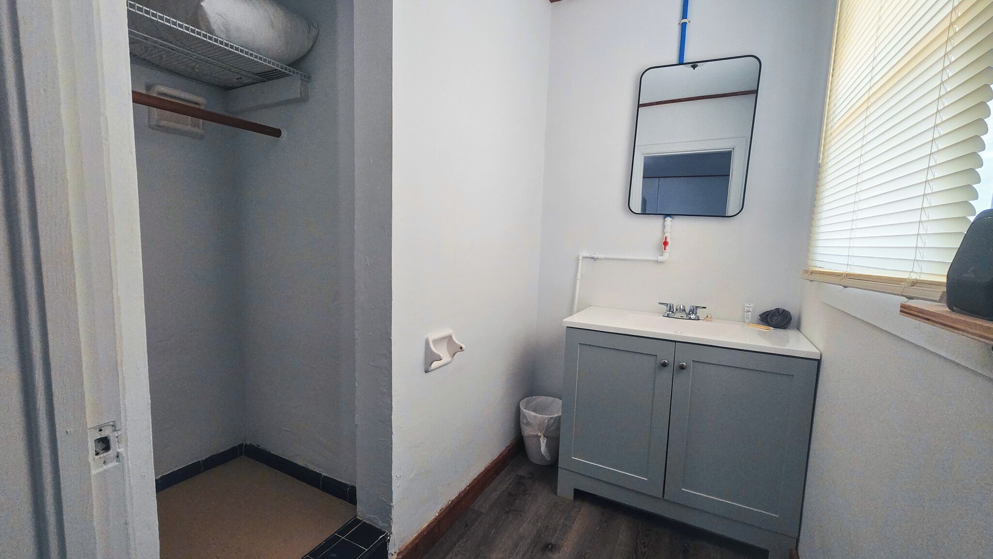 A bathroom with a white sink, mirror, and cabinet, a shower area, and a window with blinds.