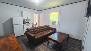 A compact living space with a kitchenette, a brown leather sofa, a wooden table, and a window with blinds.