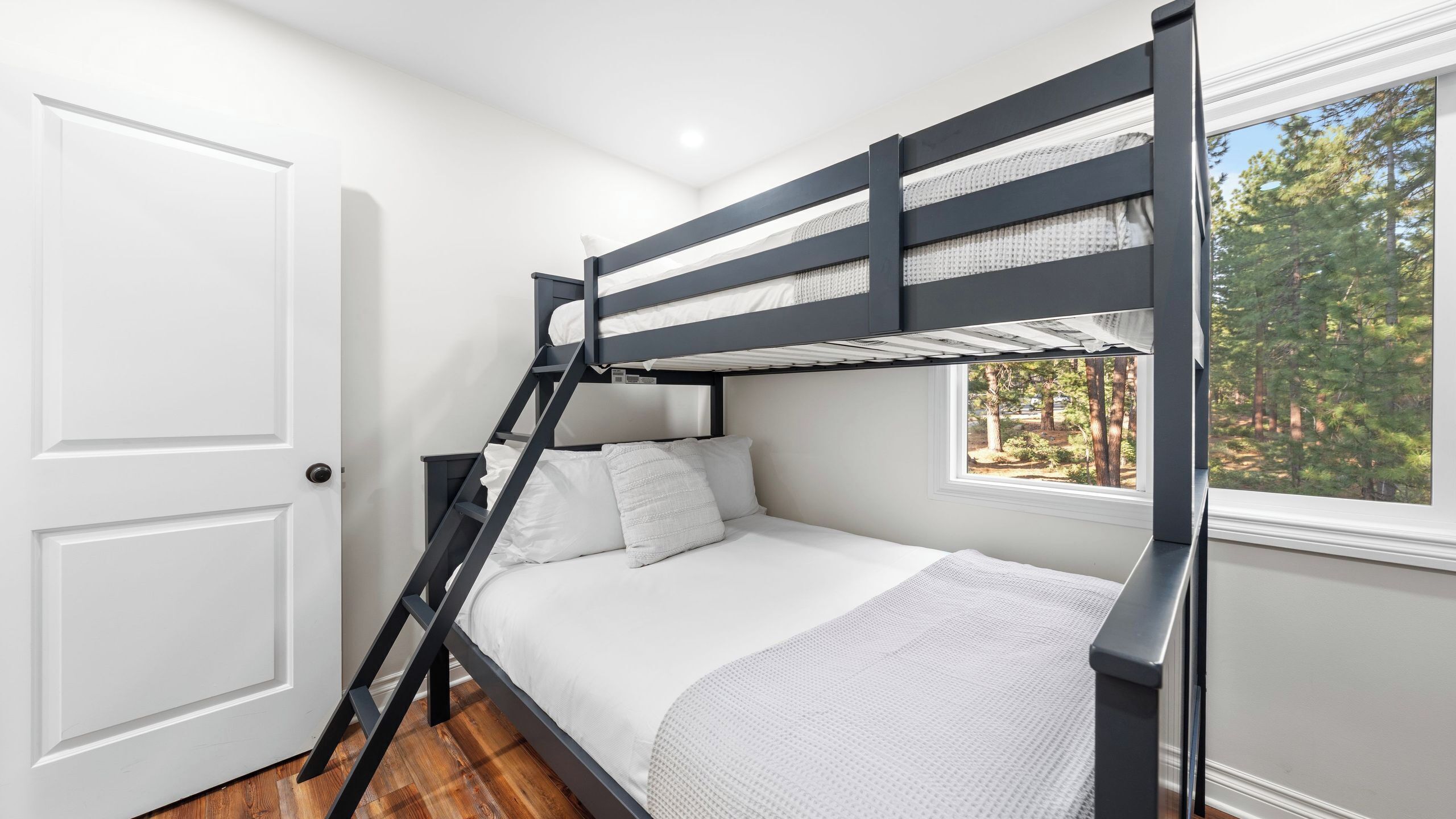 A bunk bed with a ladder, white bedding, a window with a forest view, and a door.
