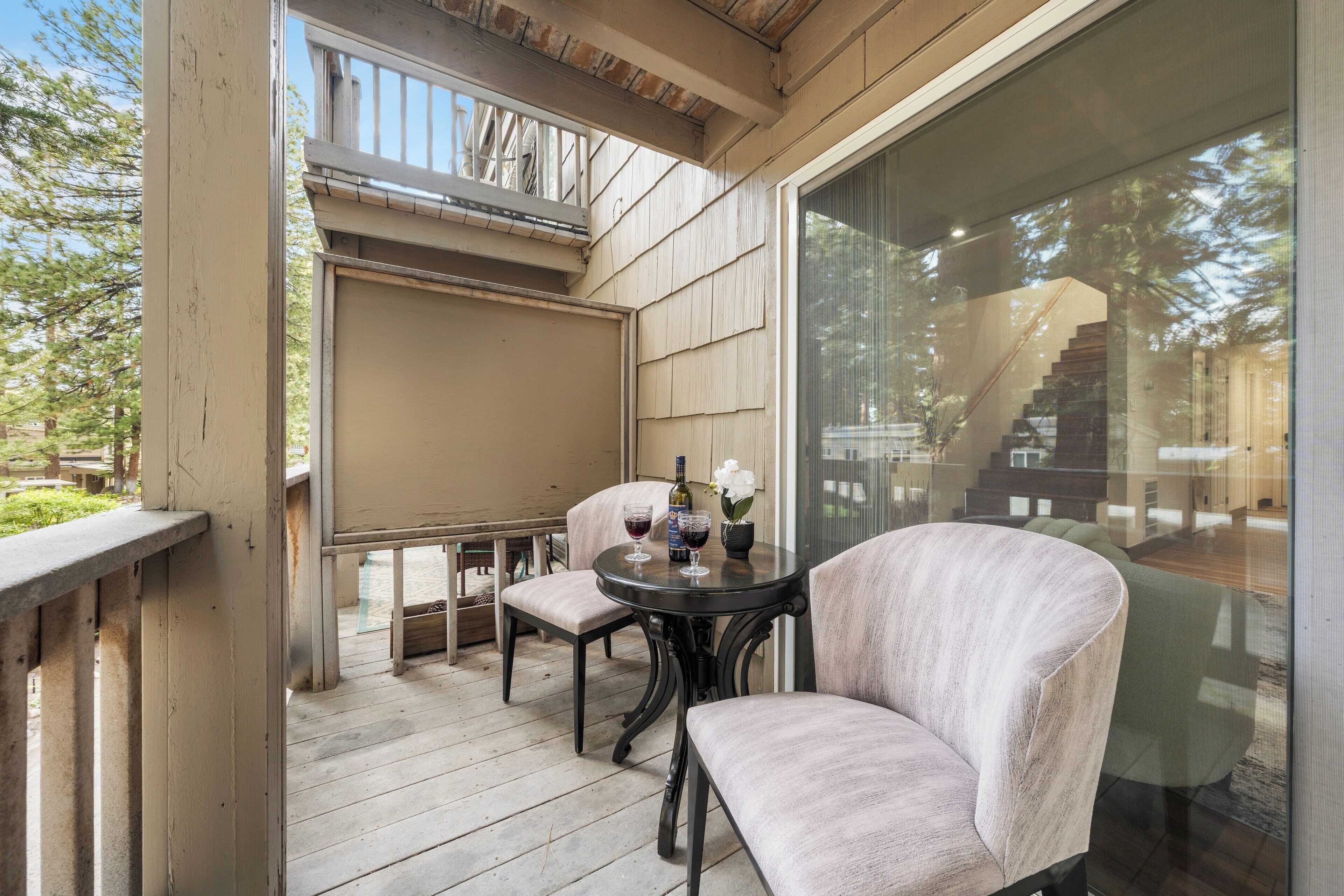 A covered outdoor seating area with a table and two chairs, a bottle of wine, and a vase with flowers.