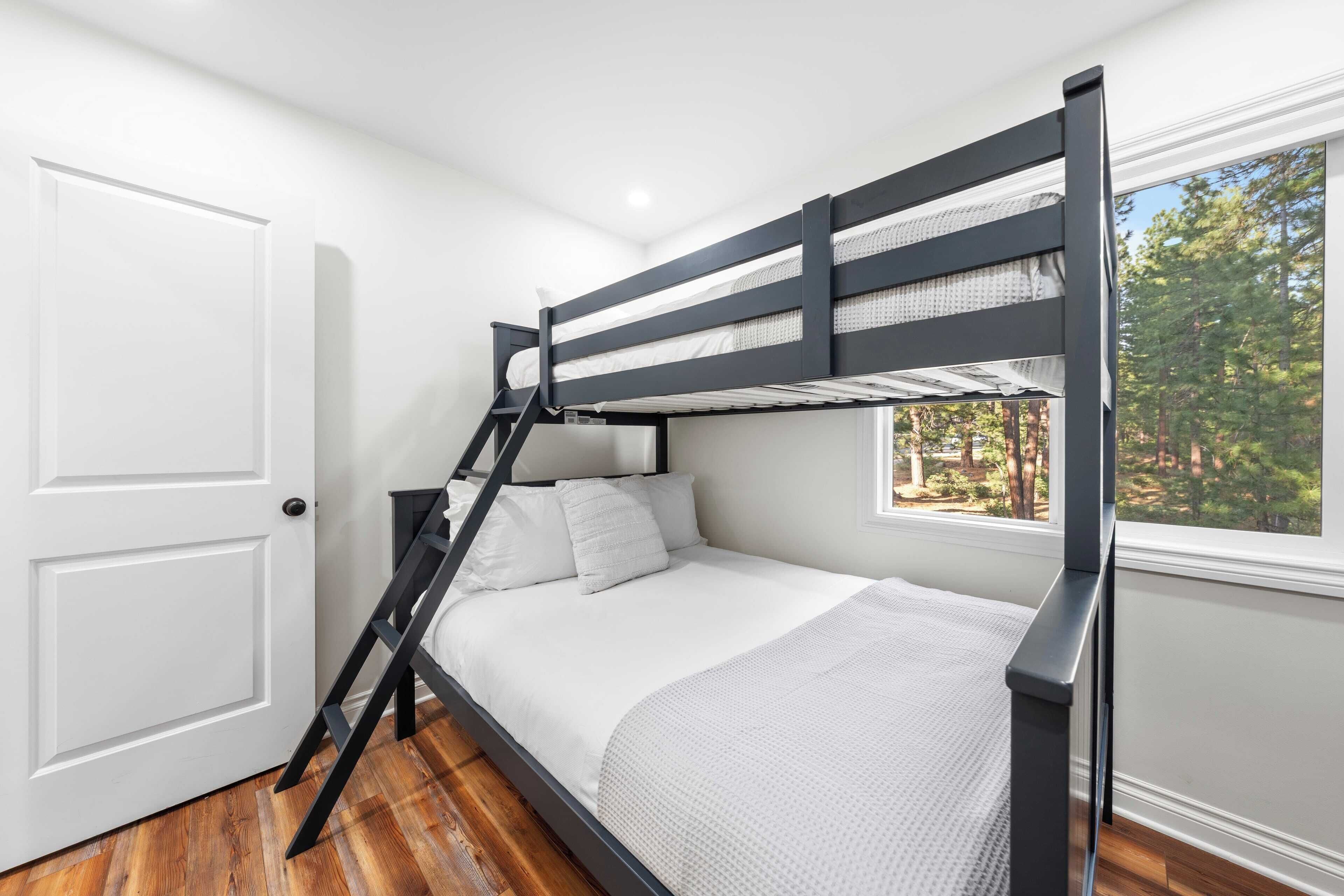 Una cama litera en una habitación con paredes blancas, suelo de madera y ventana con vistas a árboles.