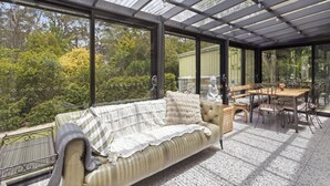 A conservatory with a cushioned sofa, dining table, and chairs, offering a view of trees and a garden. - The Orchard - Cosy A-Frame Mountain Hideaway (Leura)