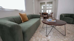 Uma sala de estar com um sofá verde, uma mesa de centro redonda de madeira e uma área de refeições ao fundo.