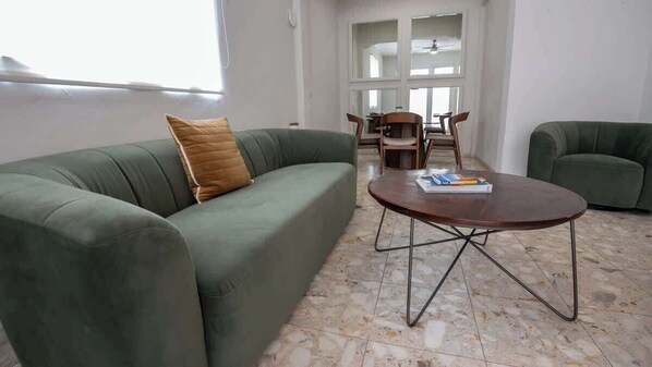 Una sala de estar con un sofá verde, una mesa de centro redonda de madera y un comedor al fondo.