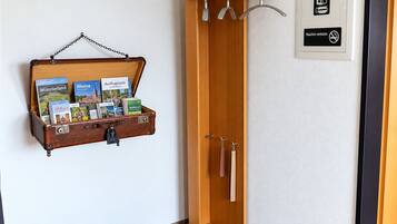 Een hotelkamer met een houten garderobe, een glazen plank met reisbrochures, een zwarte mat bij de ingang en een rookvrij sign op de muur.