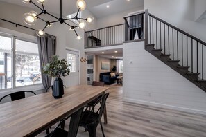 Un comedor amplio con una mesa y sillas de madera, una lámpara moderna de araña y una escalera que sube a un nivel superior.