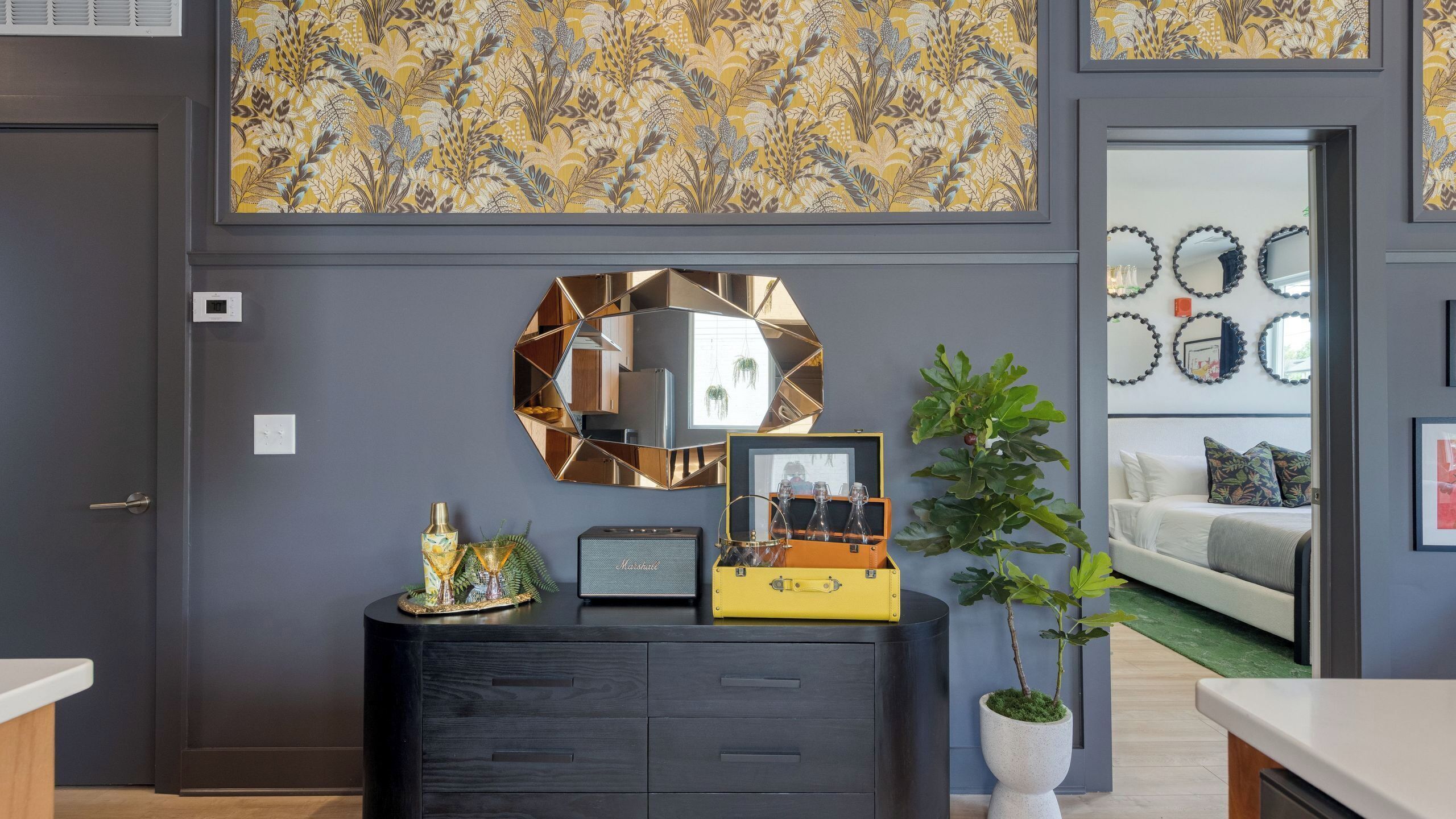 A modern living room with a dark gray wall, a decorative mirror, a yellow cabinet with glassware, and a large plant.