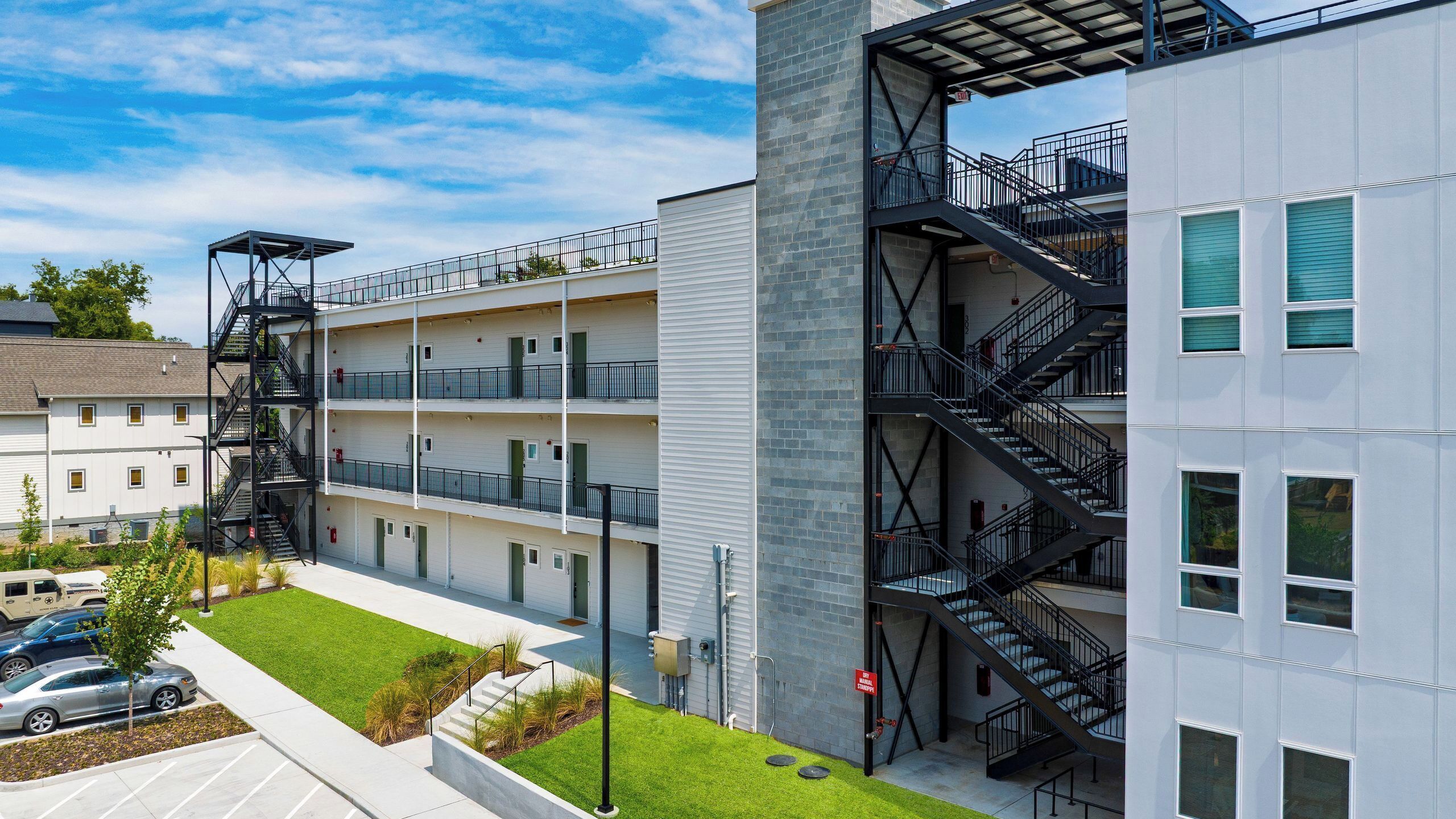 Un edificio de varios pisos con escaleras de emergencia exteriores, un estacionamiento y un patio ajardinado.