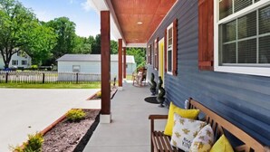 Une véranda abritée avec un banc et des plantes en pot, un ciel dégagé et un quartier résidentiel avec une clôture et des arbres.
