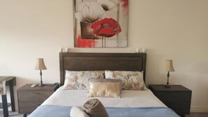 Una cama bien tendida con una manta azul, dos almohadas y una toalla enrollada sobre un cabecero de madera. Hay dos mesitas de noche con lámparas y una obra de arte colgada en la pared sobre la cama.