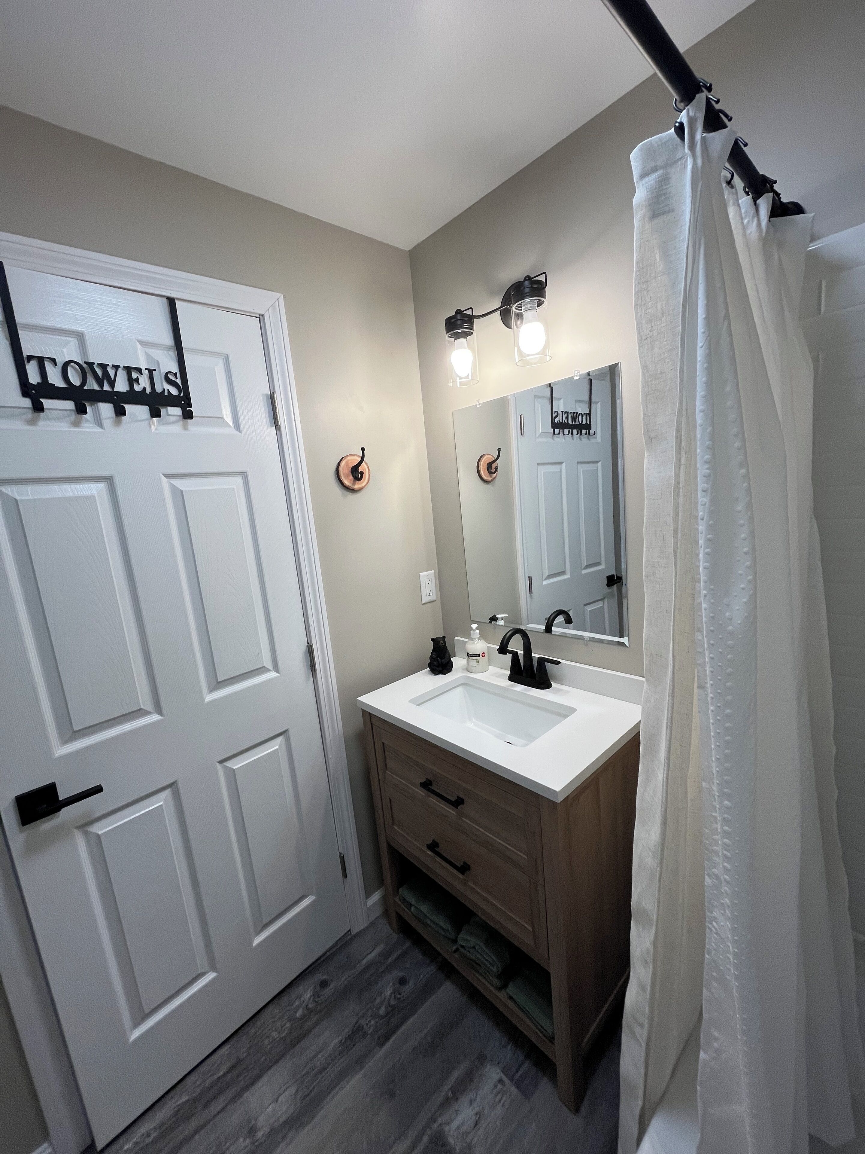A bathroom with a white sink, wooden vanity, and a mirror above it. There is a towel rack on the door and a shower curtain.