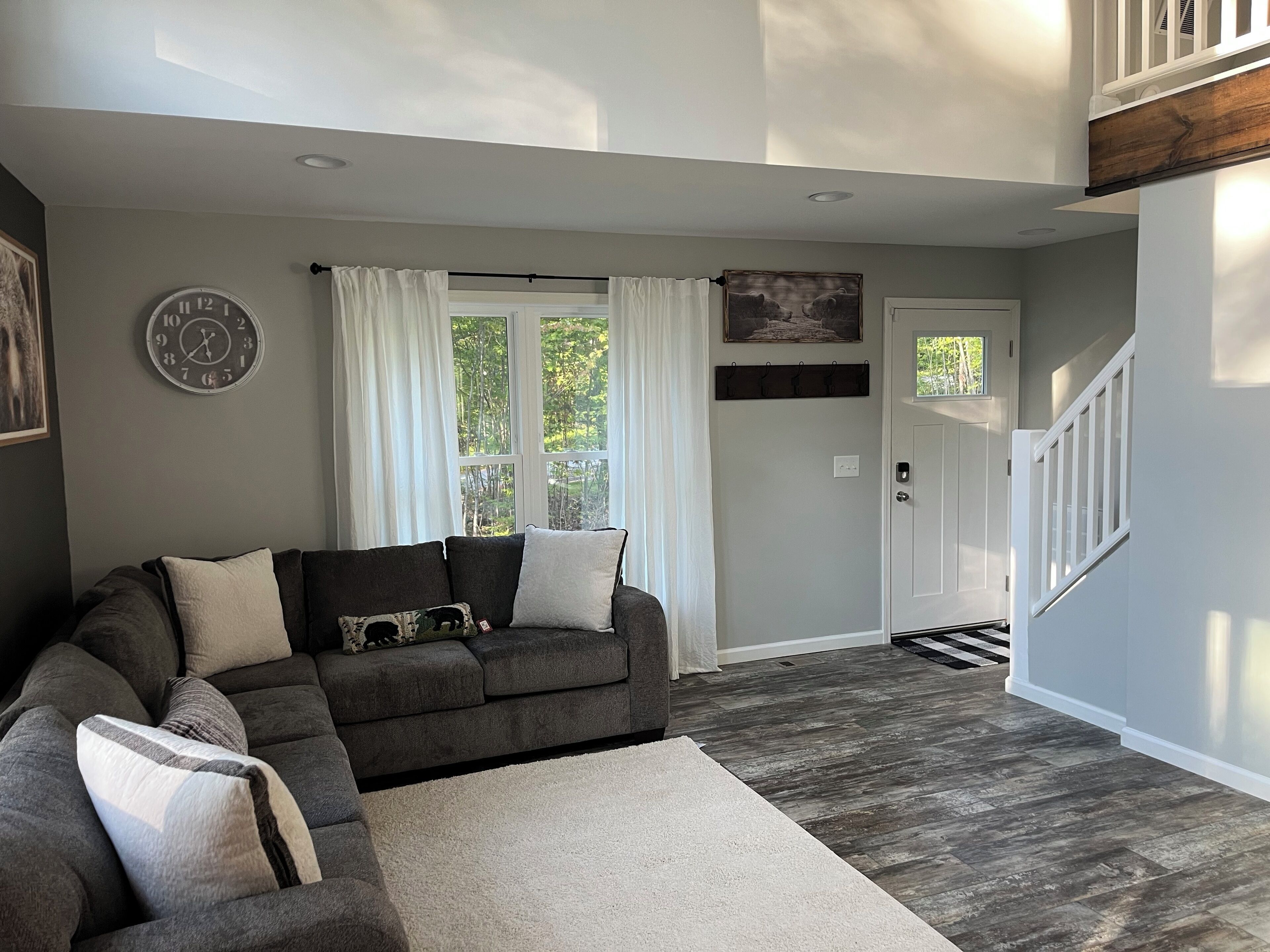 A living room with a dark grey sectional sofa, a white rug, a clock on the wall, and a door leading to a staircase.
