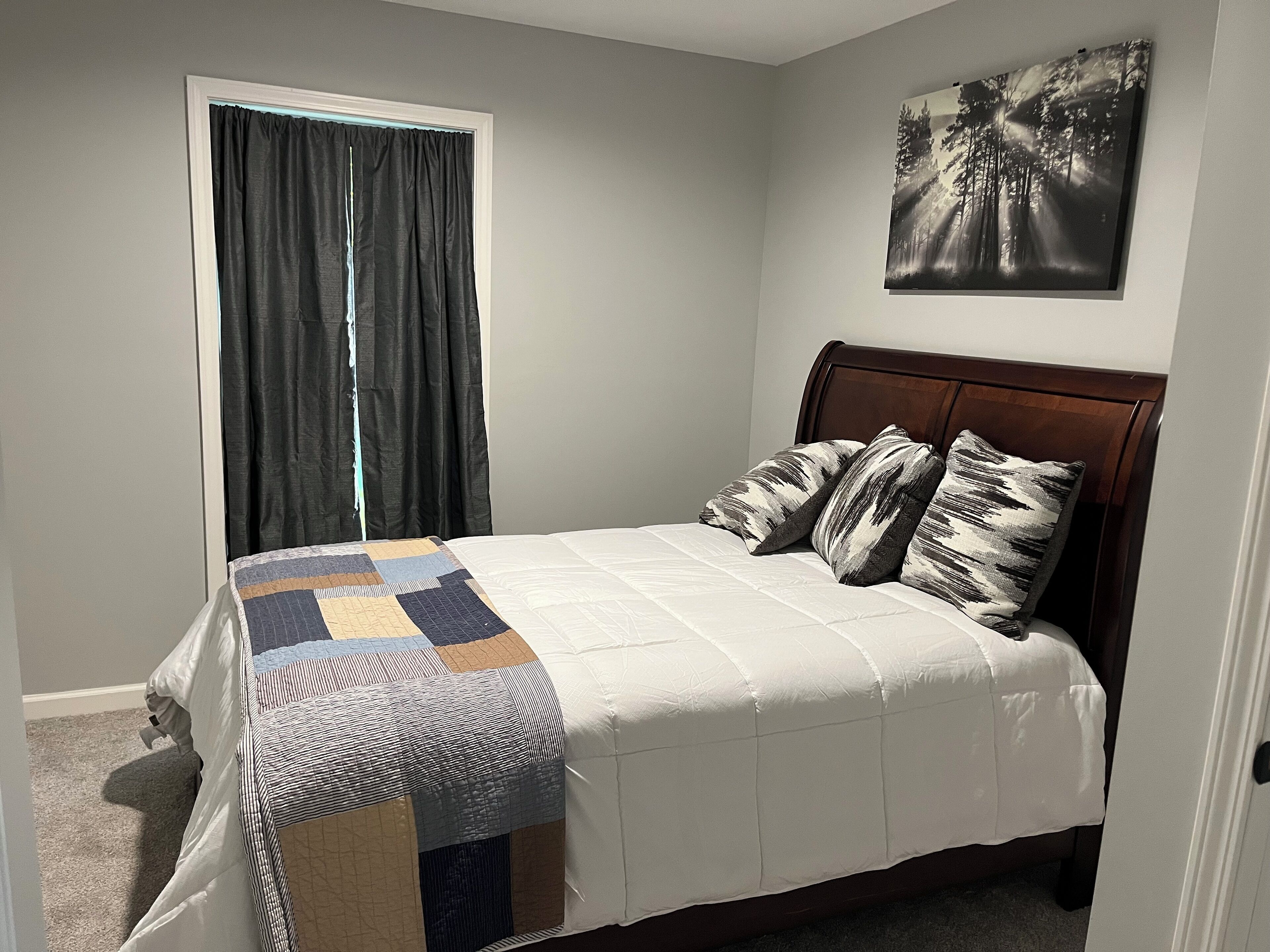 A bedroom with a bed, a window with curtains, a painting on the wall, and a carpeted floor.