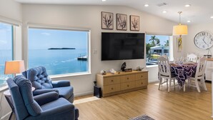 Uma sala de estar moderna com uma janela grande, uma televisão de tela plana, um espaço de jantar com mesa posta para dois e vista para o mar.
