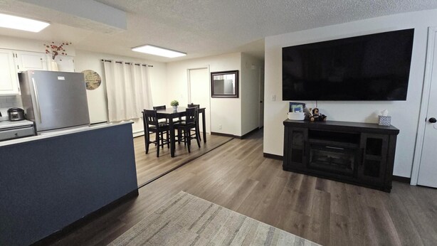A modern kitchen with a refrigerator, dining table, and chairs. There is a flat-screen TV mounted on the wall and a small cabinet below it.
