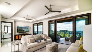 A modern living room with a sofa, coffee table, and two side tables. There are ceiling fans, a flat-screen TV, and a large window with a view of a coastal landscape. - The Only 7-Bedroom at Splendor – Luxury Oceanview Two-Level Condo with Rooftop (Playa Flamingo)