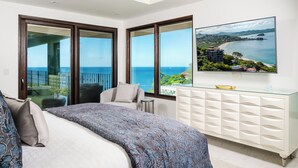 A bedroom with a large bed, a glass cabinet, and a view of the ocean through large windows. - The Only 7-Bedroom at Splendor – Luxury Oceanview Two-Level Condo with Rooftop (Playa Flamingo)