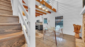 Une cuisine moderne avec des meubles blancs, un coin repas doté de chaises transparentes et un escalier en bois.