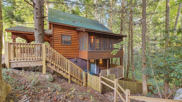 Une cabane en rondins dotée d’une terrasse en bois et d’un porche couvert, entourée d’arbres.