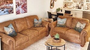 A living room with a brown leather sectional sofa, a round coffee table, and a rug. There are two framed pictures on the wall, a kitchen area with white cabinets, and a dining table with chairs.
