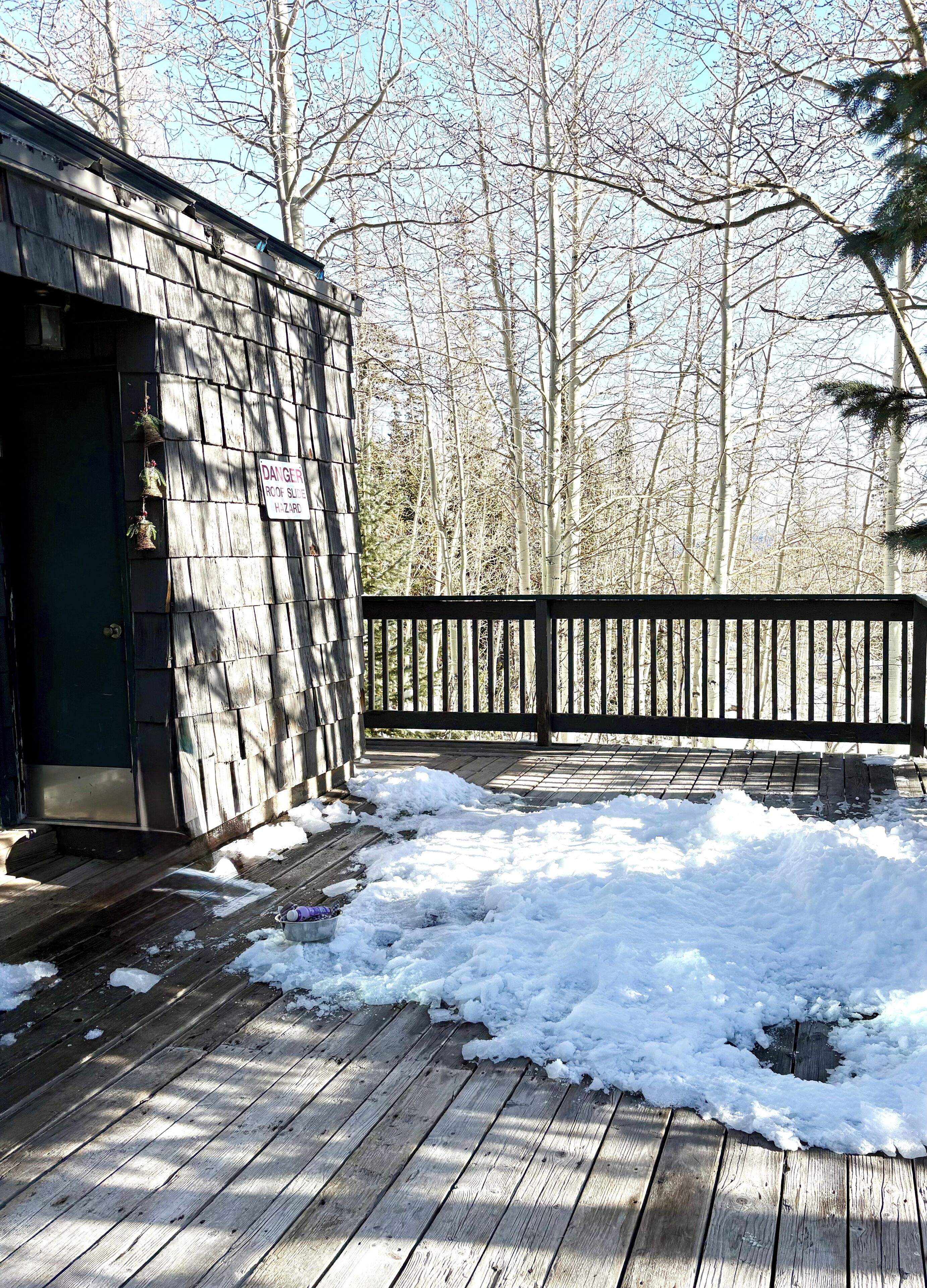 Een houten terras met sneeuw op de grond, een zwart hek en een gebouw met een waarschuwingsbord: 'DANGER ROOF SURFACE HAZARD'.