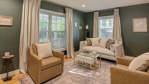Una sala de estar con un sofá color beige, un sillón marrón y una mesa de centro. Hay dos ventanas con cortinas, una lámpara de pie y cuadros en las paredes.