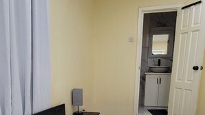 Ein ordentlich bezogenes Bett mit weißen Leinen und zwei aufgerollten Handtüchern, ein Nachttisch mit einer Lampe und Blick in einen Küchenbereich durch eine geöffnete Tür.