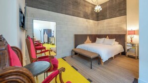 A hotel room with a bed, a bench, a TV, a mirror, and a dining area with chairs.