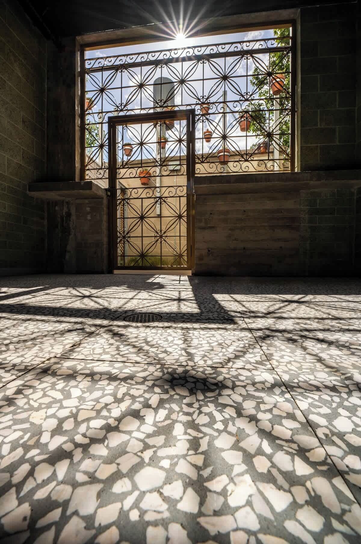 Ein mit geometrischen Mustern gefliester Boden, ein Metallgitter mit Verzierungen und ein Blick auf ein Gebäude mit Terrakotta-Dachziegeln durch das Gitter hindurch.
