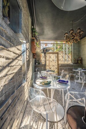 Un espace extérieur aménagé pour les repas, avec des chaises et des tables en plastique transparent, un portail en fer forgé et un lustre.