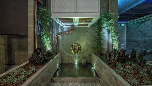 A modern indoor space with a central reflective sculpture, surrounded by planters and a metal staircase.