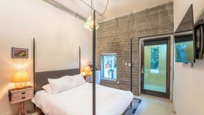 A modern hotel room with a large bed, a nightstand with lamps, a sliding glass door to a balcony, and a mounted TV.