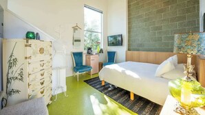 A modern bedroom with a large bed, a dresser, a desk, and a chair. There is a window providing natural light and a view of greenery outside.