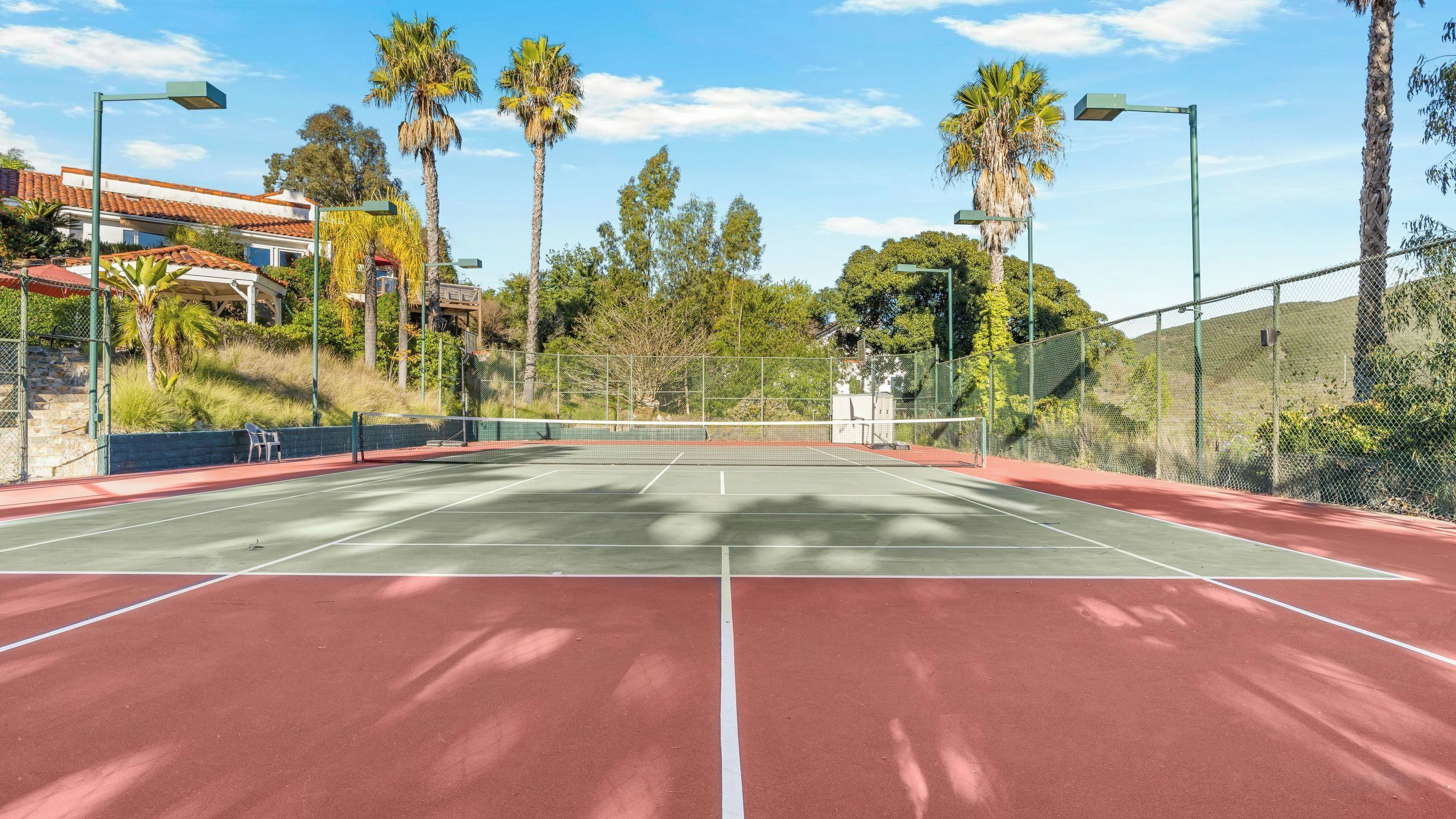 En udendørs tennisbane med rød overflade, omgivet af hegn, palmer og en klar himmel.