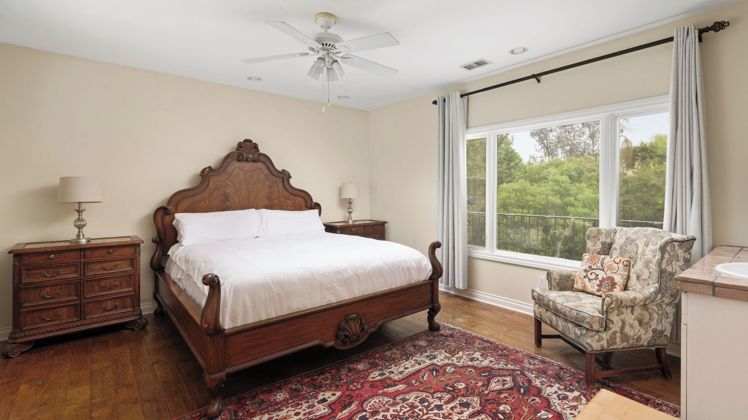 A bedroom with a large bed, a chair, a nightstand, a ceiling fan, and a window with a view.