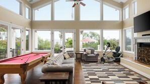 A spacious room with a pool table, a sofa, exercise equipment, and large windows offering a view of the outdoors.