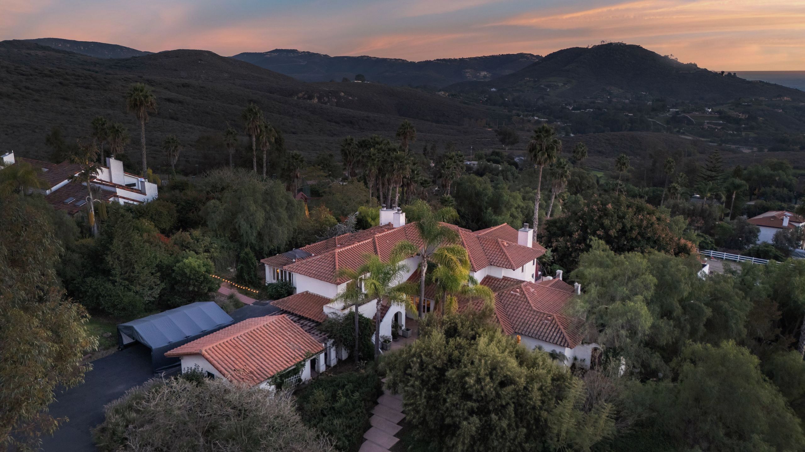 A residential area with houses featuring red-tiled roofs, surrounded by greenery and hills in the background.