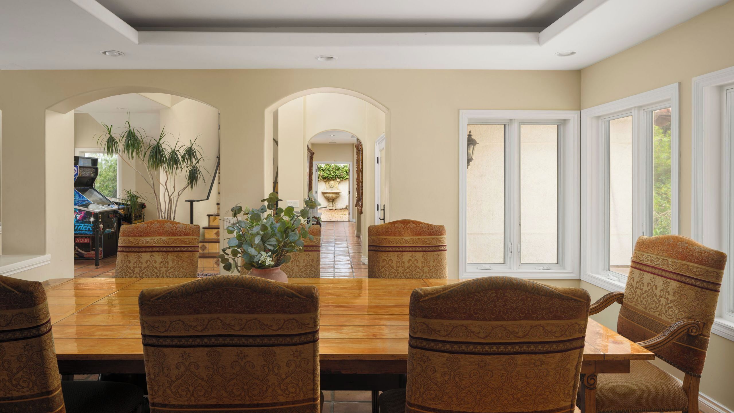 A dining room with a large wooden table, patterned chairs, and a view through an archway to a garden.