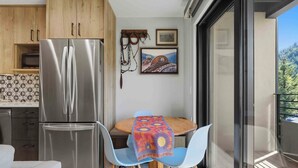 Een kleine keuken met een roestvrijstalen koelkast, een houten eettafel met een kleurrijke tafelkleed en blauwe stoelen.