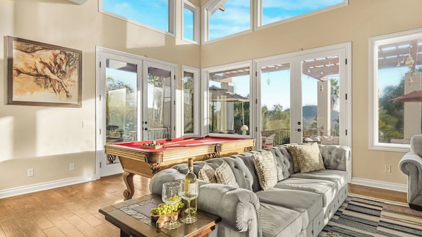 A spacious living area with a pool table, comfortable seating, and large windows offering views of the outdoors.