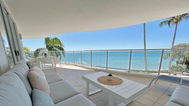A sunlit outdoor seating area with a view of the sea, featuring a sofa, a table, and a railing. - Amazing Water View Apartment - Pool, Gym, Parking. (Urangan)