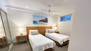 A hotel room with two single beds, a ceiling fan, a painting of a beach, and a window with a view. - Amazing Water View Apartment - Pool, Gym, Parking. (Urangan)