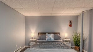 Une chambre à coucher avec un grand lit, deux tables de chevet avec des lampes, une table de nuit, une œuvre d’art accrochée au mur et une plante en pot.