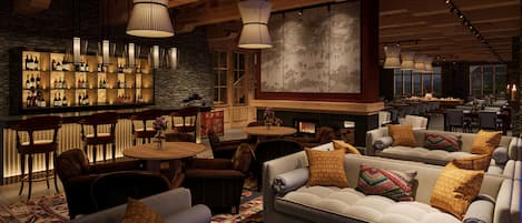 A modern bar area with wooden beams, a stone wall, and a large painting. There are multiple seating areas with sofas and chairs, and a bar stocked with bottles.