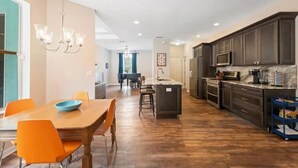 Une cuisine spacieuse avec des meubles de couleur sombre, un plan de travail en marbre et un coin repas agrémenté de chaises orange et d’une table en bois.
