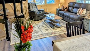 A cozy cabin interior with a loft bed, a sofa, a dining table, and a chair. - Bar Harbor Cabin with Treehouse Suite & Hot Tub (Hancock)