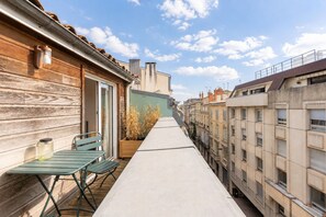 A narrow outdoor balcony with a wooden wall on one side and a building on the other. There is a metal table and two chairs, and a potted plant is visible. - Bright Apartment in the City Centre, Terrace, A/C (Bordeaux)