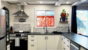 Una cocina moderna con armarios blancos, una campana extractora de acero inoxidable, horno empotrado, pileta doble y una ventana con cortinas rojas.