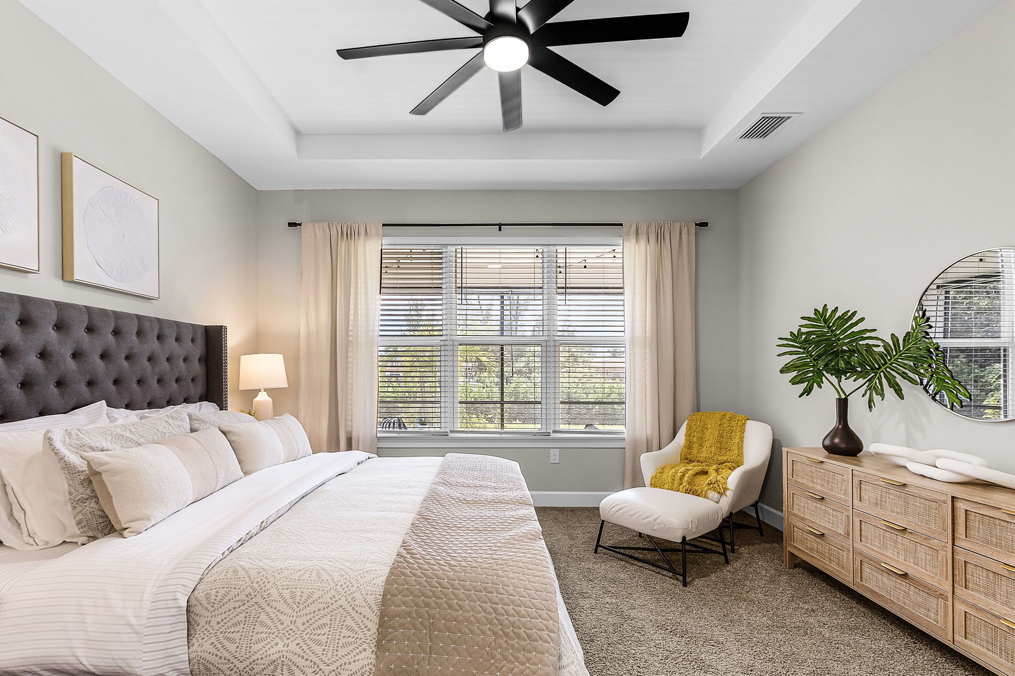 A bedroom with a large bed, a ceiling fan, a window with blinds, a chair, a bedside table, and a mirror.