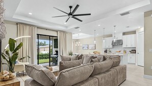 Un salon moderne avec un grand canapé gris, un ventilateur de plafond et une vue sur le jardin, accessible par une porte-fenêtre coulissante.